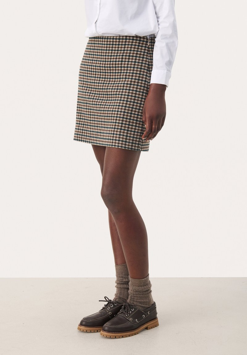Femme portant une jupe à carreaux marron et beige, des chaussettes marron, des chaussures noires à lacets et une chemise blanche à manches longues, debout de profil.