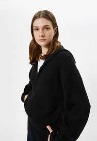 Schwarzer Strickpullover mit Reißverschluss am Hals, oversized Schnitt und strukturierter Oberfläche. Hat eine Kapuze und lange Ärmel mit gerippten Bündchen.