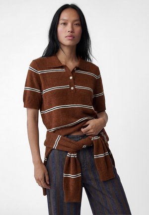 Donna con capelli lunghi e neri che indossa una polo a maniche corte marrone a righe e un maglione abbinato legato in vita a pantaloni a righe blu, in piedi davanti a uno sfondo bianco.