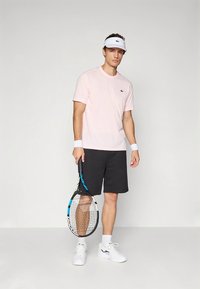 Lys rosa kortermet skjorte, svarte shorts, hvit visir, håndleddsbånd og hvite joggesko. Holder et tennisracket med blå detaljer.