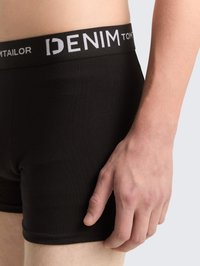 Närbild av en person som bär svarta boxerkalsonger med en märkesmidja märkt "DENIM TOM TAILOR" mot en neutral bakgrund.