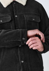 Chaqueta de pana negra con un collar de piel sintética blanca, que presenta bolsillos frontales y botones a presión en los puños y el pecho. Rayas verticales texturizadas.