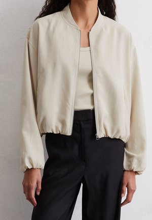 Chaqueta bomber crema con cremallera y puños elásticos, llevada sobre un top de canalé y pantalones negros de talle alto, en una persona de pie.