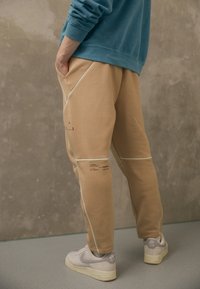 Beige tepláky s bílými švy a potištěnými detaily, spárované se šedými teniskami. Materiál vypadá měkce a neformálně.