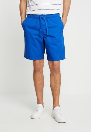 Niebieskie bawełniane szorty z elastycznym pasem, troczkiem i bocznymi kieszeniami. Tkanina wydaje się lekka i gładka. Stylizowane z białymi tenisówkami.