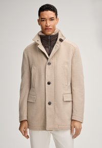 Beige wolmix jas met een hoge kraag, dubbele knoopsluiting, twee voorkruiden en een gewatteerde bruine binnenlaag.