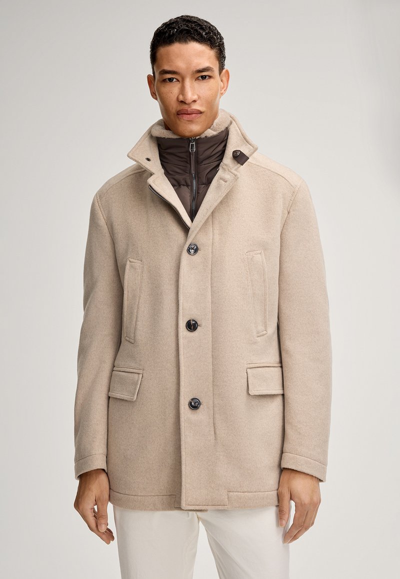 Cappotto in misto lana beige con colletto alto, chiusura a doppio bottone, due tasche frontali e un rivestimento interno in brown trapuntato.