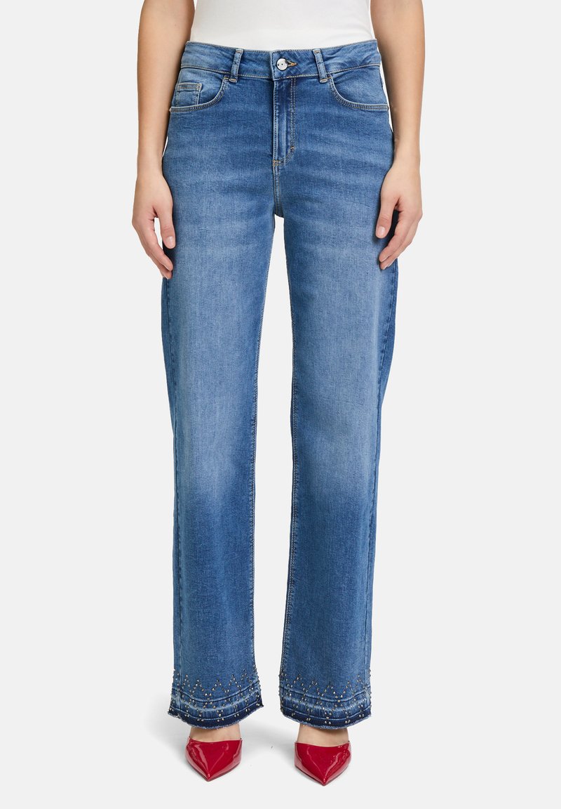 Jean large en denim bleu clair avec taille haute, ourlet orné et décoloration subtile, associés à des escarpins rouges pointus.