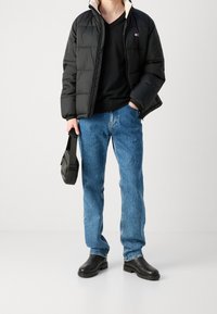 Doudoune noire avec doublure beige, pull noir à col en V, jeans en denim bleu et bottes noires. Comprend un sac bandoulière noir.
