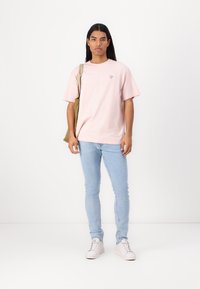 Camiseta de algodón de color rosa claro con mangas cortas y un pequeño logo. Combinada con jeans skinny de color azul claro y zapatillas blancas. Bolsa de lona al hombro.