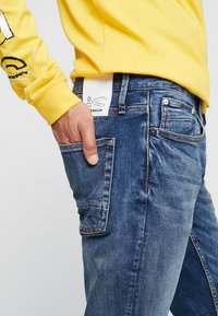 Jeans i denim i en mellanblå tvätt, med fram- och bakficka, slitna detaljer och en synlig skötseletikett vid midjebandet.