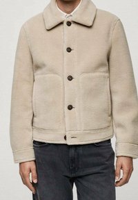 Beige Fleecejacke mit großen Taschen und dunklen Knöpfen, über einem weißen Hemd mit Kragen getragen, kombiniert mit schwarzen Jeans.