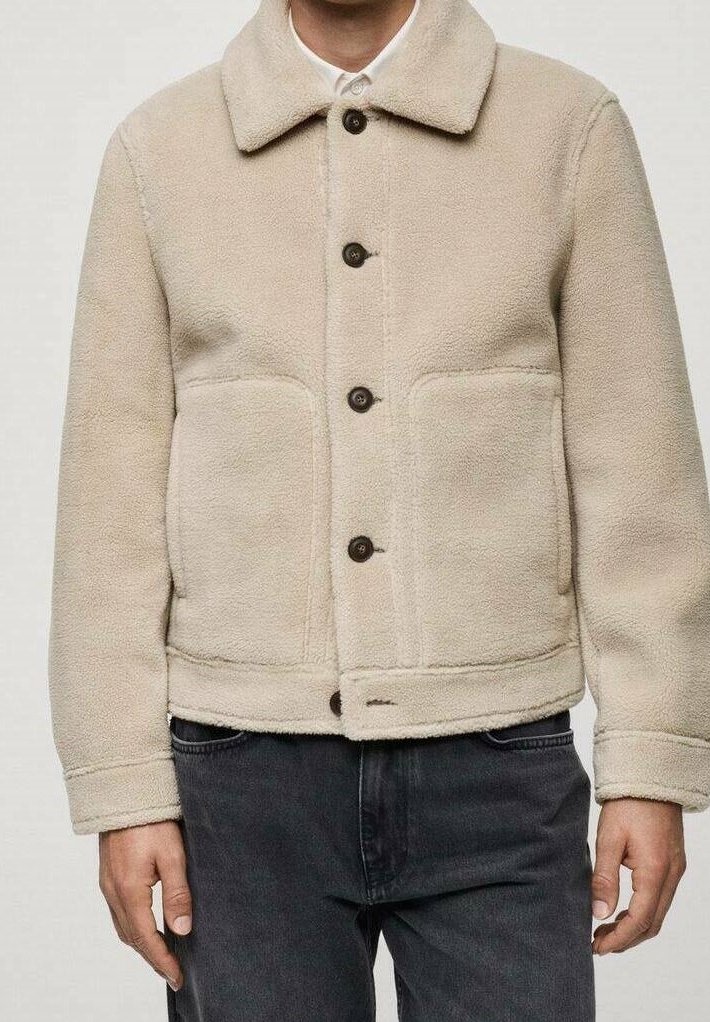 Beige Fleecejacke mit großen Taschen und dunklen Knöpfen, über einem weißen Hemd mit Kragen getragen, kombiniert mit schwarzen Jeans.