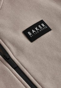 Vaaleanbeige vetoketjullinen fleece, jossa on musta suorakulmainen etiketti, jossa lukee "BAKER by TED BAKER" ja sileä, pehmeä rakenne.