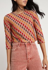 Femme portant un pull court texturé avec des motifs losanges rouges, jaunes et bruns et un jean taille haute rose délavé.