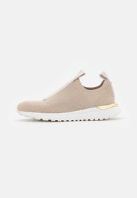 Sneaker beige senza lacci con tomaia in rete lavorata a maglia, suola bianca e dettaglio dorato sul tallone. Presenta una superficie testurizzata e un design minimalista.