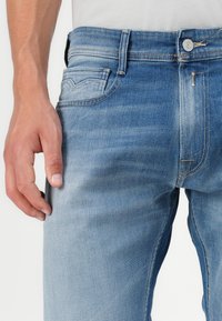Light blue denim jeans with a slim fit, five pockets, and subtle fading. Features a metal button closure and reinforced stitching details.