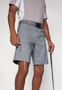 Calças cinzentas com uma textura suave, bolsos frontais, um bolso lateral com fecho e um cinto preto, combinadas com uma camisa cinza e branca.