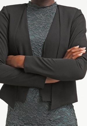 Osoba o ciemnej karnacji, ubrana w czarną marynarkę i zieloną koronkową sukienkę, z rękami skrzyżowanymi na piersi.