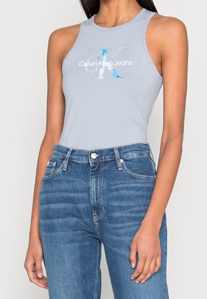 Kobieta ubrana w jasnoniebieski, bezrękawnik Calvin Klein Jeans oraz niebieskie dżinsy z wysokim stanem, stojąca na tle prostego, jasnego tła.