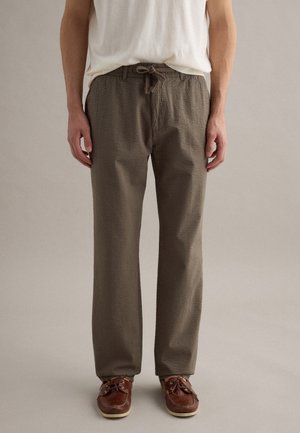 Man draagt textuur bruine broek met trekkoord, bruine leren veterschoenen en een ingestopt wit T-shirt, staand tegen een neutrale achtergrond.