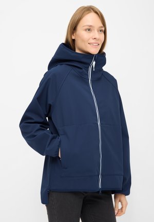 Mujer sonriente con chaqueta azul marino con capucha y cremallera blanca, con las manos en los bolsillos, de pie frente a un fondo claro liso.