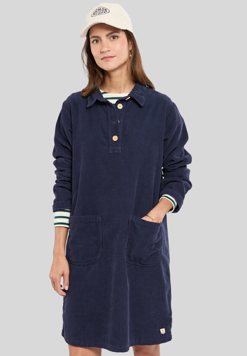 Robe en velours côtelé bleu marine avec un col, trois boutons, des manches longues, deux poches avant et des poignets rayés. Étiquette logo notable à l'ourlet.
