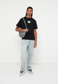Camiseta negra con un parche gráfico colorido, pantalones vaqueros holgados de color azul claro, bolso cruzado gris y zapatillas gris-azul. Diseño sencillo y ajuste informal.