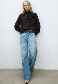 Blusa negra con un detalle de lazada, combinada con jeans de pierna ancha de color azul claro deslavado y un cinturón marrón. Un calzado sencillo complementa el conjunto.