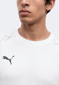 Νεαρός άνδρας με λευκό μπλουζάκι Puma και μαύρο λογότυπο Puma στο στήθος, εμφανίζεται από το πηγούνι έως το επάνω μέρος του στήθους σε μονόχρωμο φόντο.