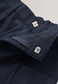 Gros plan sur un pantalon en tissu foncé avec une braguette ouverte et une fermeture à pression sur la ceinture.