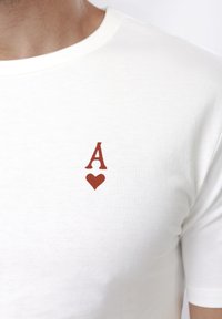 Λευκό μπλουζάκι με ένα μικρό κόκκινο γράμμα "Α" πάνω από ένα κόκκινο σύμβολο καρδιάς στο αριστερό στήθος, φορεμένο από ένα άτομο.