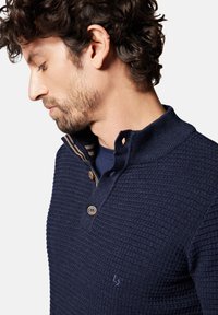 Dzianinowa granatowa koszulka polo z teksturowanym wzorem, z dwoma guzikami i ściągaczem przy kołnierzyku. Z przodu znajduje się subtelne logo.