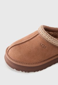 Brown suede UGG slip-on slipper with stitched trim and soft lining, displayed on a white background.