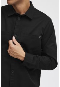 Black long-sleeve shirt with a button-down collar, two chest pockets, and textured fabric, featuring black buttons and subtle branding detail.