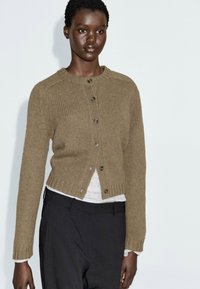 Khaki knoop-up cardigan gemaakt van een gestructureerde brei, met een ronde hals, lange mouwen en geribbelde manchetten, gelaagd over een witte top.