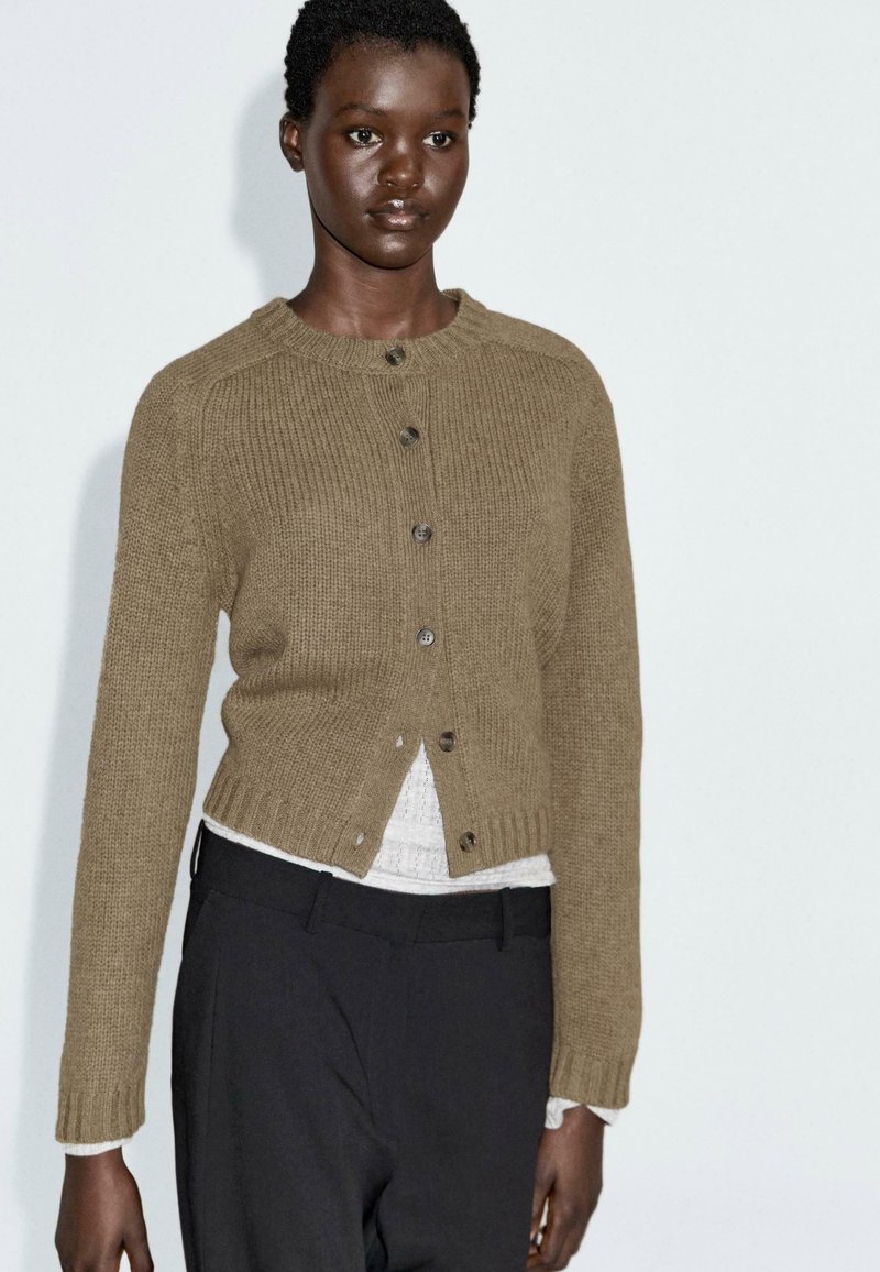 Khaki knoop-up cardigan gemaakt van een gestructureerde brei, met een ronde hals, lange mouwen en geribbelde manchetten, gelaagd over een witte top.