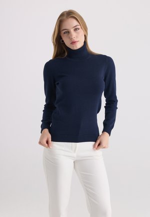 Femme aux longs cheveux blonds portant un pull à col roulé bleu marine et un pantalon blanc, debout devant un fond clair uni.