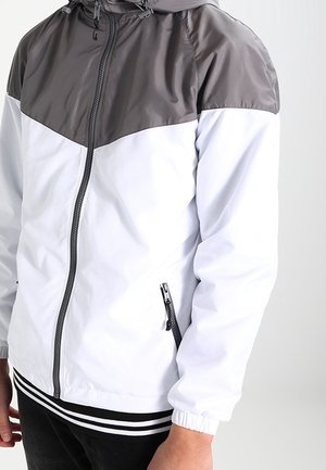 Mand iført en hvid og grå vindjakke med lynlås, elastiske manchetter og lynlåslomme på siden, stående foran en ensfarvet baggrund.