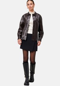 Veste chemise en simili cuir brun brillant, à boutons sur le devant, col évasé. Sous-vêtement en col roulé blanc, jupe portefeuille bleu marine, bottes noires jusqu'aux genoux.