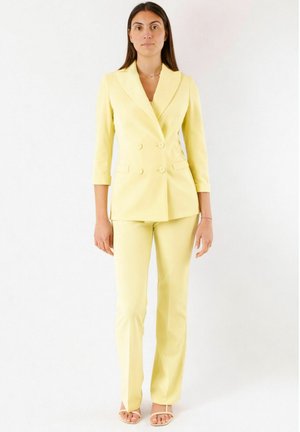 Donna in piedi, indossa un blazer doppiopetto giallo chiaro con pantaloni abbinati e sandali scoperti, su uno sfondo bianco uniforme.