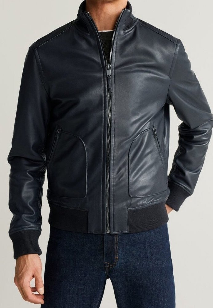 Homme portant une veste en cuir noire zippée avec poignets et ourlet côtelés, associée à un jean bleu foncé sur un fond uni.