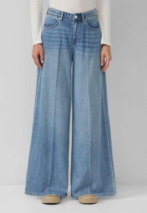 Femme portant un jean denim bleu clair à jambes larges et des chaussures beiges, debout devant un fond gris uni.