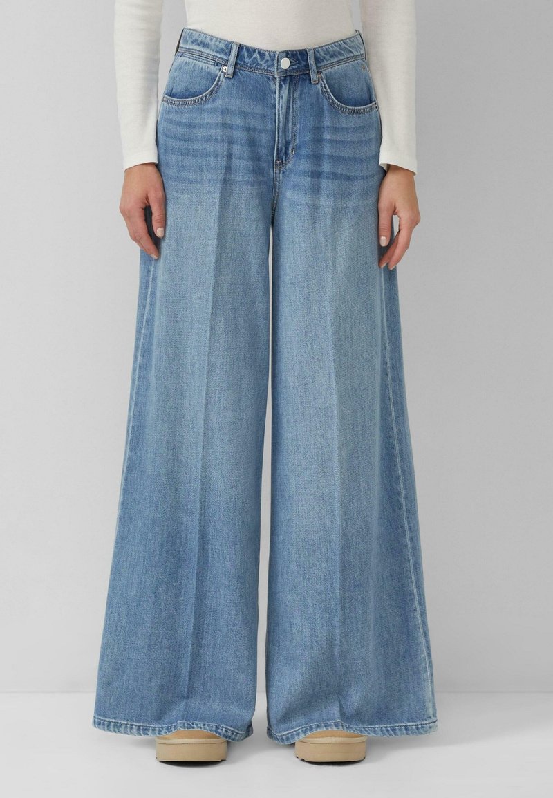 Femme portant un jean denim bleu clair à jambes larges et des chaussures beiges, debout devant un fond gris uni.
