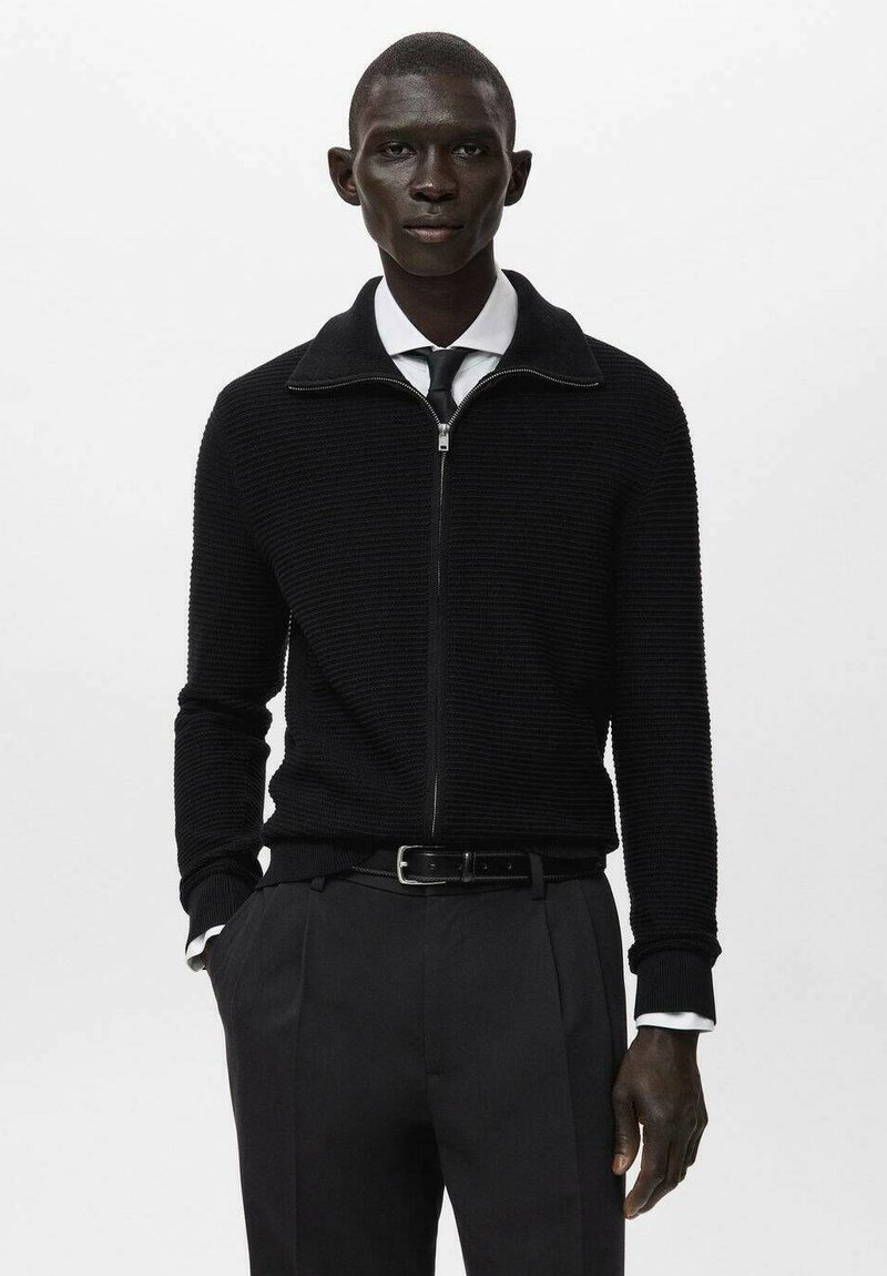 Czarny sweter zapinany na zamek, o fakturowanym wzorze dzianiny, z kołnierzykiem i długimi rękawami, zestawiony z białą koszulą i czarnymi spodniami.