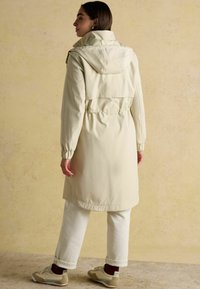 Sieviete, kas ģērbusies garā bēšā kapucīna mētelī, baltās biksēs un bēšos sneakers, stāv pretim bēšai teksturētai fona, skatoties prom.