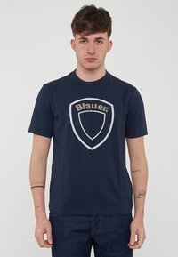 Marineblå t-shirt lavet af bomuld med korte ærmer, der har et stort, lyst skjoldlogo med teksten "Blauer." Trykt design foran.