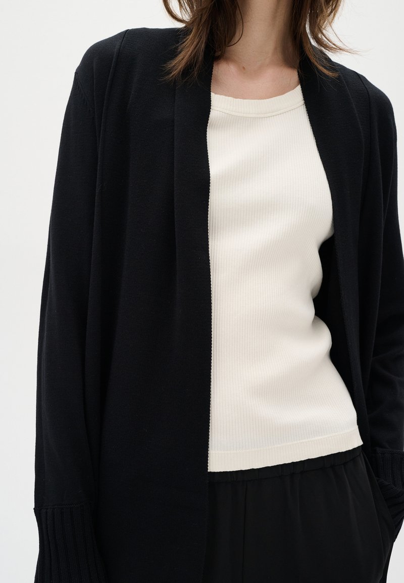 Schwarze offene Strickjacke mit gerippten Bündchen über einem cremefarbenen RIP-Top. Glatte Textur, lockerer Schnitt, ergänzt durch lose schwarze Hose.