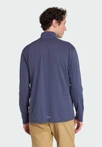 Námornícka dlhá rukávová košeľa s vysokým golierom, hladkou textúrou a detaily šitia. Logo na spodnom leme. Nosená s béžovými šortkami.