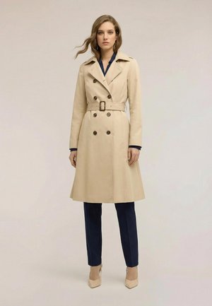 Vrouw met beige trenchcoat met riem, gecombineerd met marineblauwe broek en hakken, staand tegen een effen lichte achtergrond.